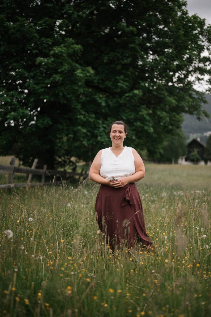 Frau im weißen Oberteil und brauem Rock steht in einer blühenden Wiese vor einem Baum.