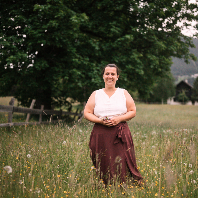 Frau im weißen Oberteil und brauem Rock steht in einer blühenden Wiese vor einem Baum.