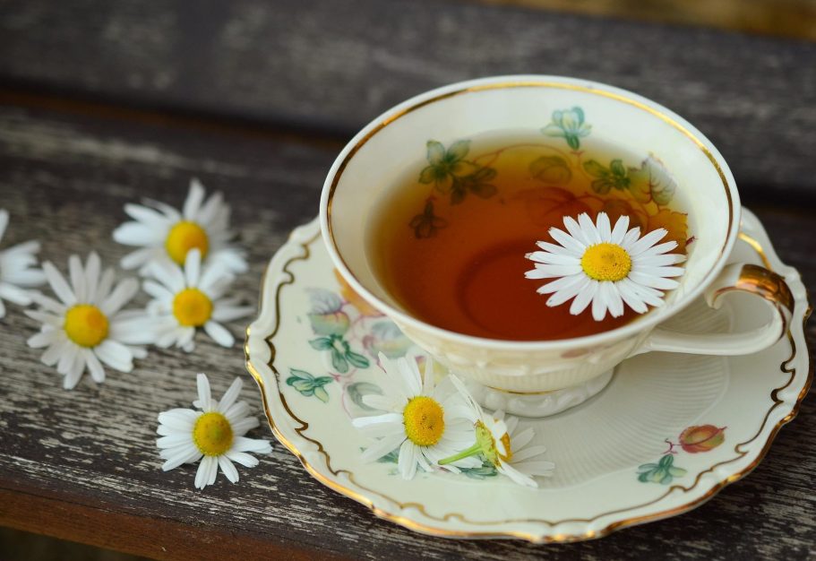 Tasse mit Gänseblümchentee und einer Blüte Gänseblume im Tee, auf einem Holzbrett, umgeben von weiteren Gänseblümchen.