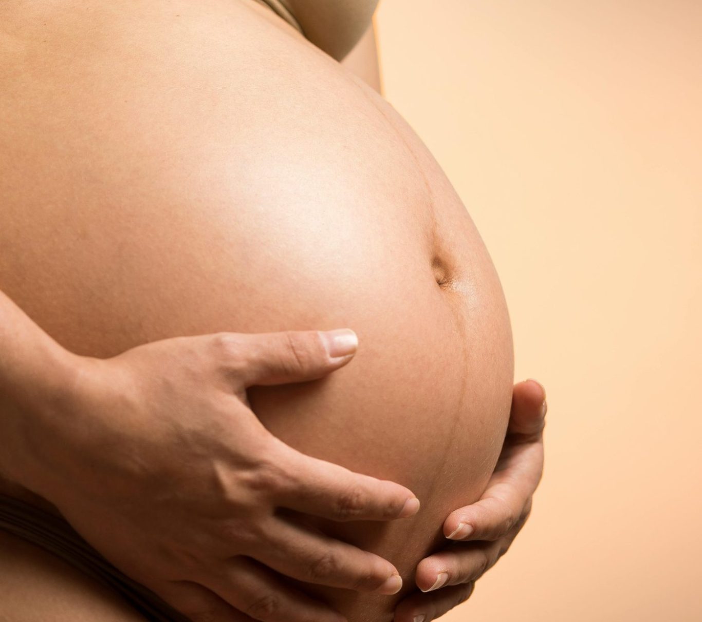 Schwangere Person hält ihren runden Babybauch mit beiden Händen fest.